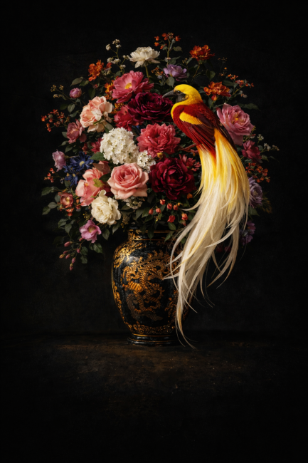 Stilleven met kleurrijke bloemen in een sierlijke vaas en een geel-rode paradijsvogel tegen een donkere achtergrond