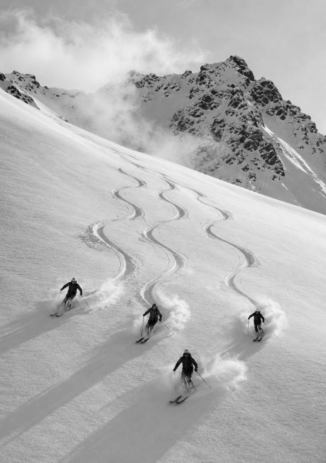 Zwart-wit wanddecoratie van vier skiërs die door verse poedersneeuw afdalen op een berghelling met indrukwekkende bergtoppen op de achtergrond.