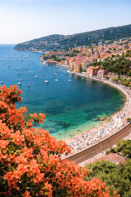 Panoramisch uitzicht over de baai van Villefranche-sur-Mer aan de Franse Rivièra met turquoise zee, zeilboten, strand en kleurrijke mediterrane huizen.